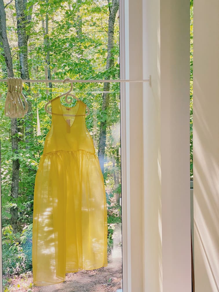 A Coco Silk Dress by Noémiah, crafted from sheer yellow silk organza, hangs on a clothesline indoors near a large window. Sunlight filters through the trees outside, creating soft shadows on the floor and walls and accentuating its relaxed fit.