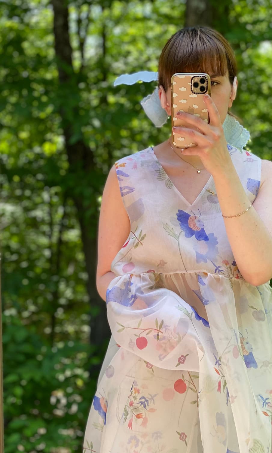 Wearing the Coco Silk Dress by Noémiah, a person takes a mirror selfie in an outdoor setting filled with vibrant greenery, their face partially hidden by the phone.