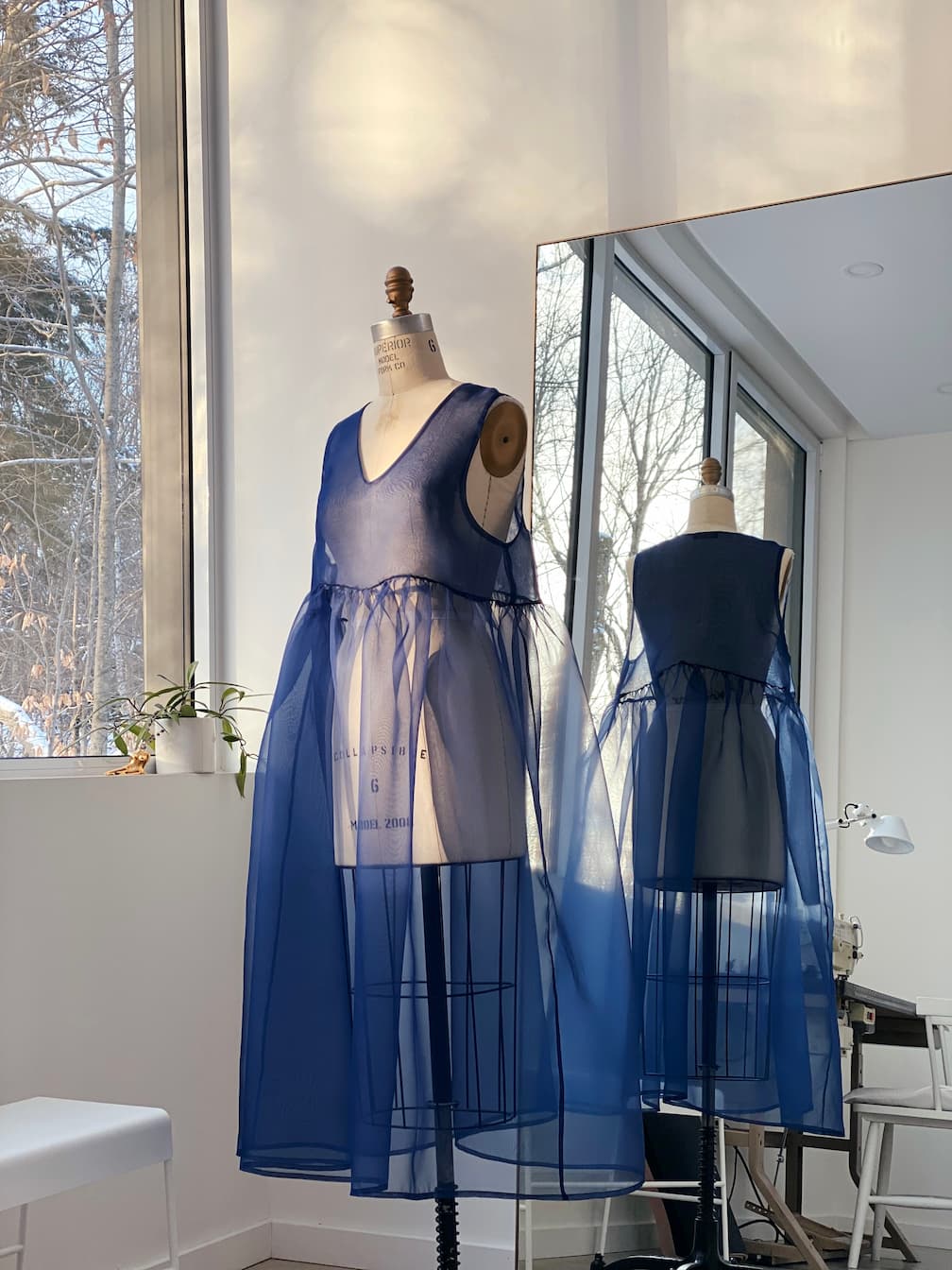 A blue sheer Noémiah Coco silk dress is displayed on a dress form in a modern room with large windows and a light interior.