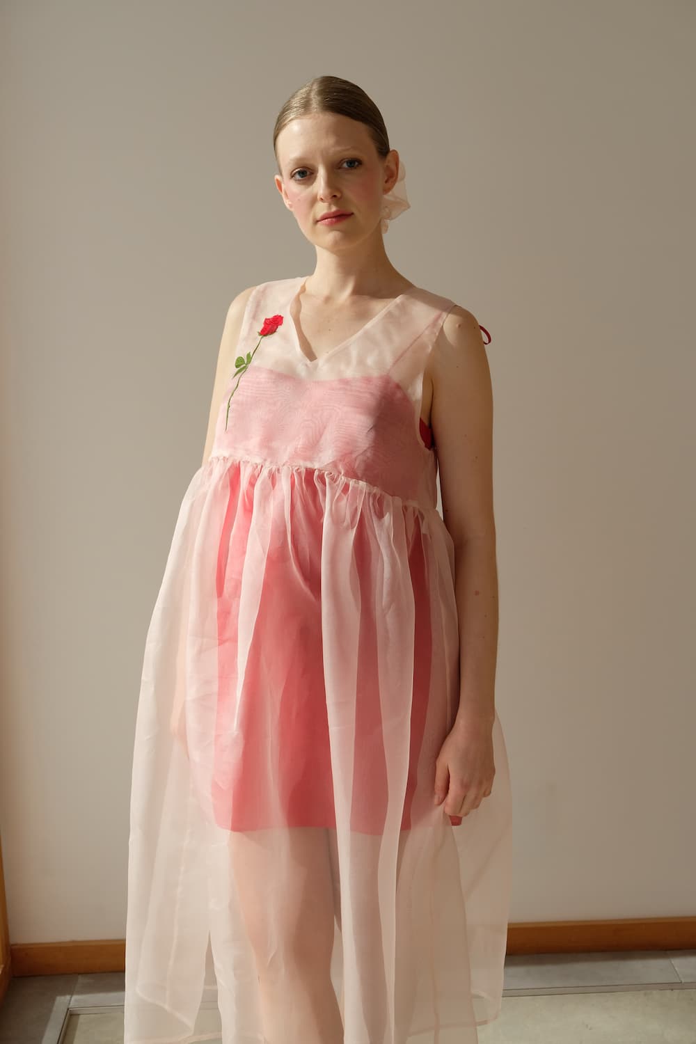 A woman poses against a plain backdrop in a luxurious Florence Silk Dress — Classic by Noémiah. This elegant pink dress boasts an A-line silhouette crafted from silk organza, with a sheer overlay and a delicate red rose accent on the shoulder; her light skin contrasts with her pulled-back hair.