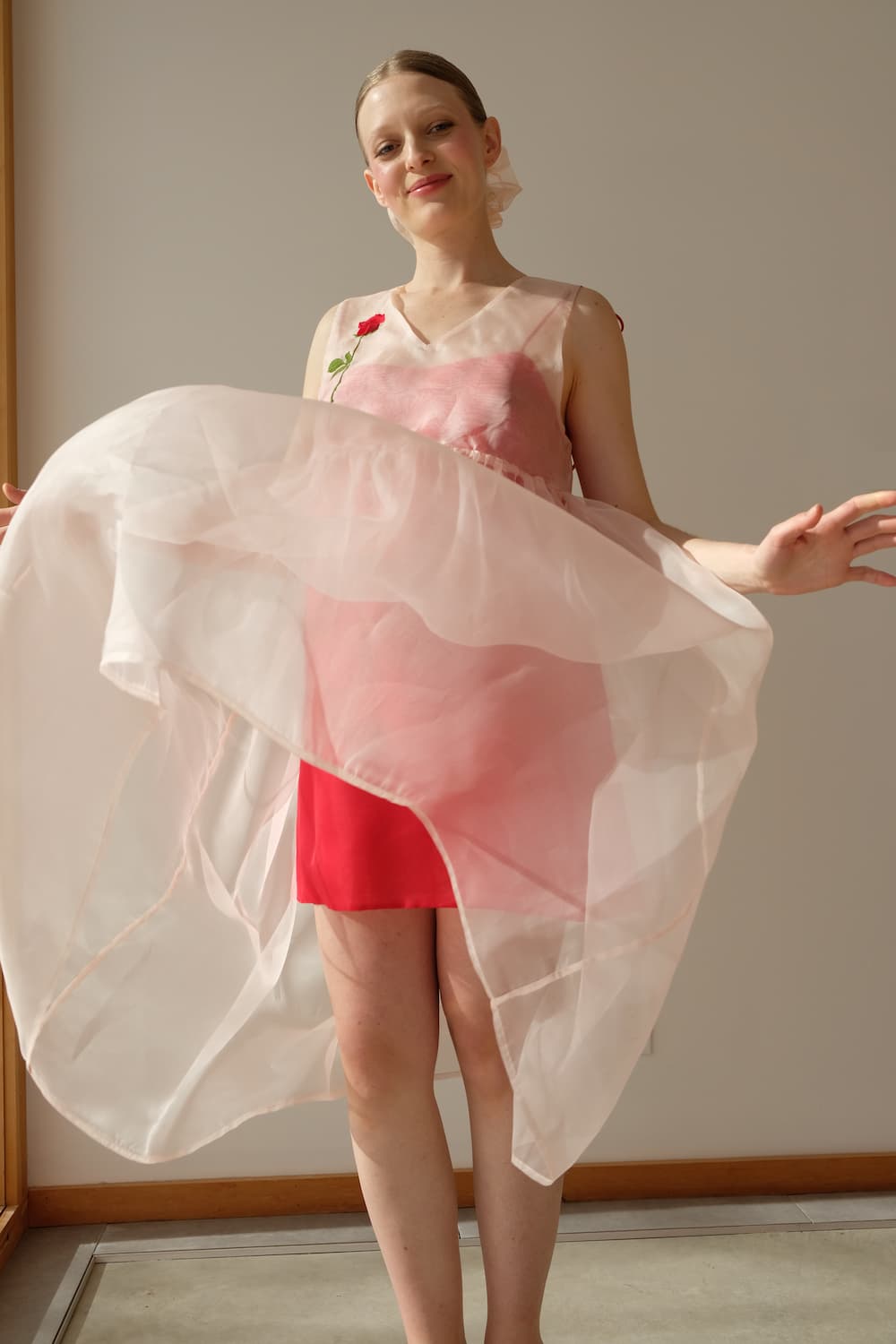 A woman stands indoors, smiling gently in a Noémiah Florence Silk Dress — Classic. The flowing pink and red organza fabric forms an A-line silhouette, accented by a rose embroidered on the shoulder, as sunlight illuminates her from the left.