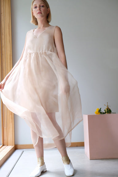 A person wearing a flowing, sheer Coco Silk Dress by Noémiah stands indoors near a wooden-framed window. They have on white chunky shoes with golden ankle socks that perfectly complement the relaxed fit of the dress. In the background, there is a pink box adorned with flowers.