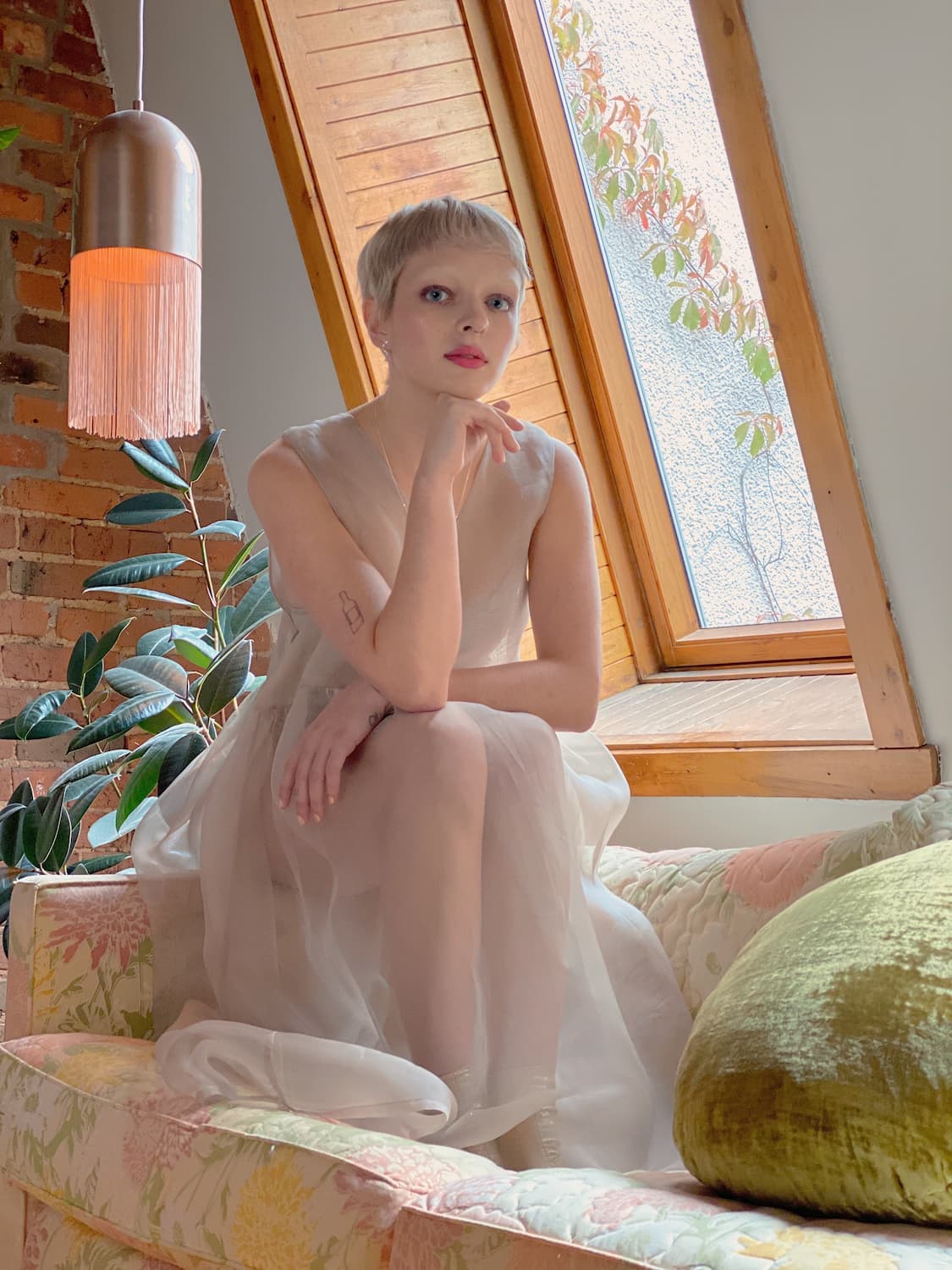 A person with short hair sits on a floral couch near a window, wearing an elegant Coco Silk Dress by Noémiah. They rest their chin on their hand, with a plant and brick wall forming the background. Soft lighting adds to the cozy ambiance.