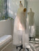 A dress form stands in a sunlit room showcasing the semi-transparent Coco Silk Dress by Noémiah. Crafted from delicate silk organza, the dress is mirrored nearby, while a potted plant rests on the windowsill next to a white bench. Soft shadows are cast as natural light filters through the window.