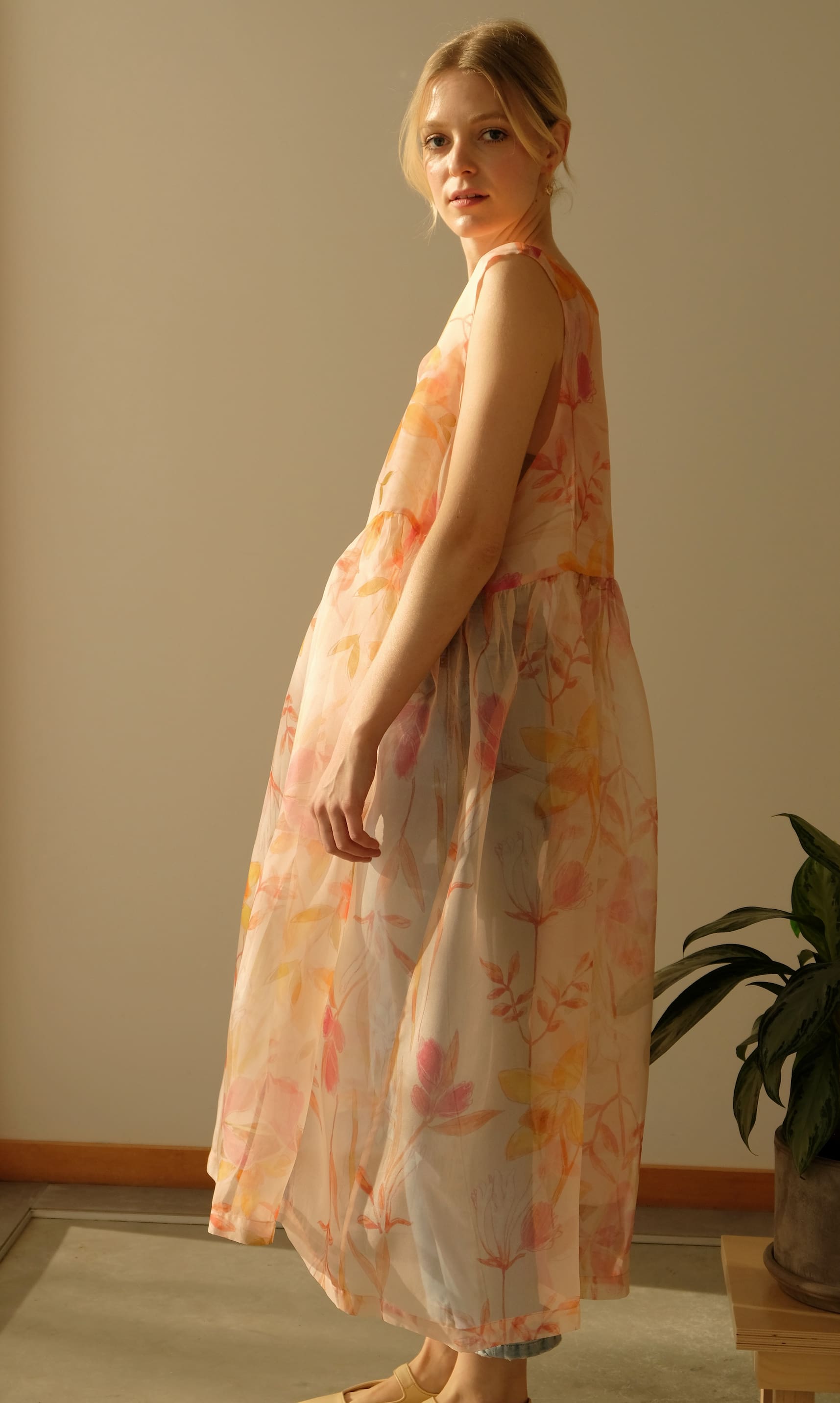 In a softly lit room, someone is wearing the Coco Silk Dress by Noémiah, featuring a relaxed fit and a flowing floral print. The semi-transparent silk organza fabric is adorned with blooms in shades of orange, pink, and yellow. A green plant can be seen partially in the background on the right.