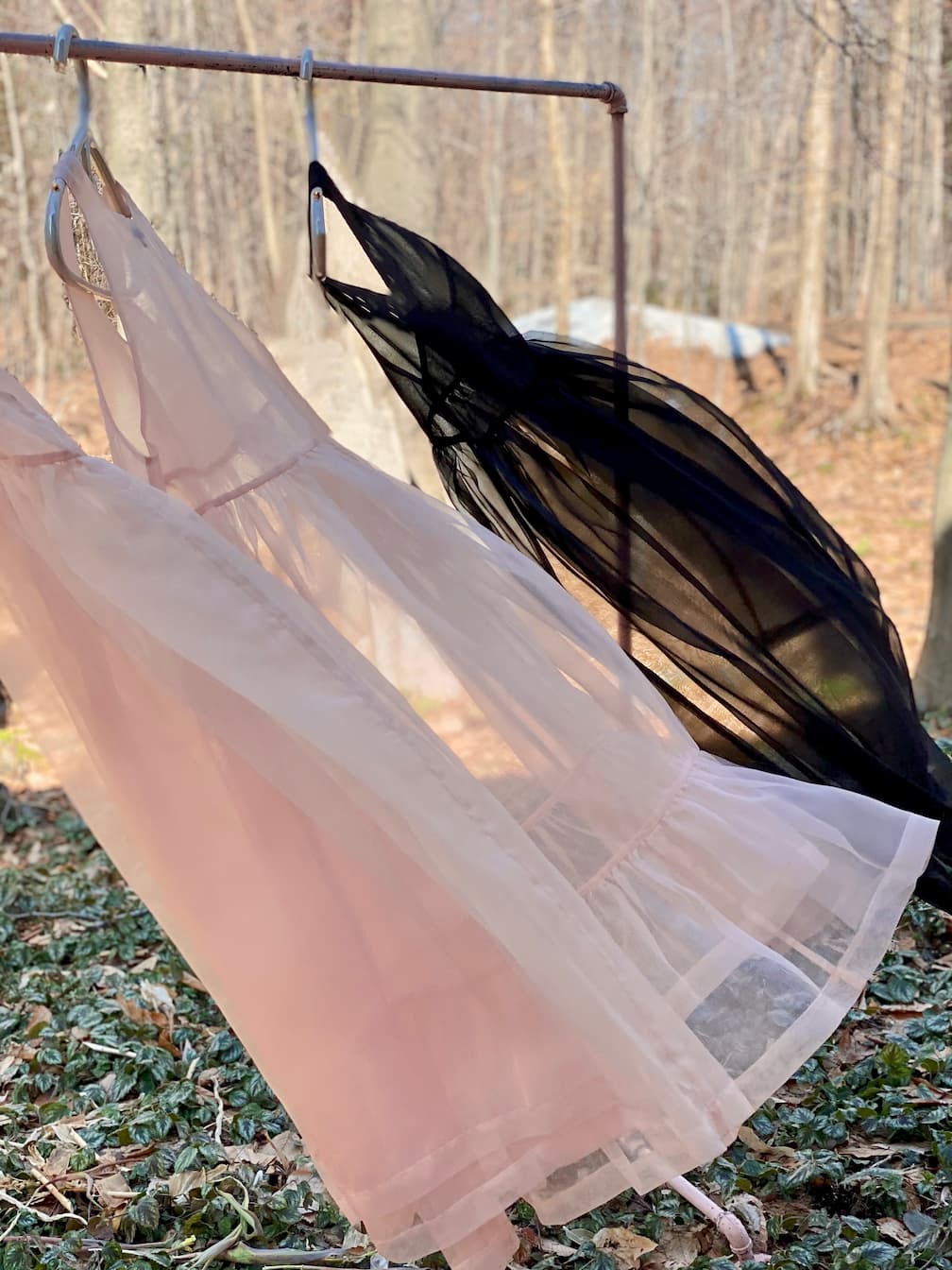 Three exquisite dresses, featuring a Noémiah Coco Silk Dress, elegantly hang on a metal rod outdoors. Two of them are in pale pink, while the third is black with a relaxed fit. The serene background showcases a wooded area with bare trees and scattered leaves.