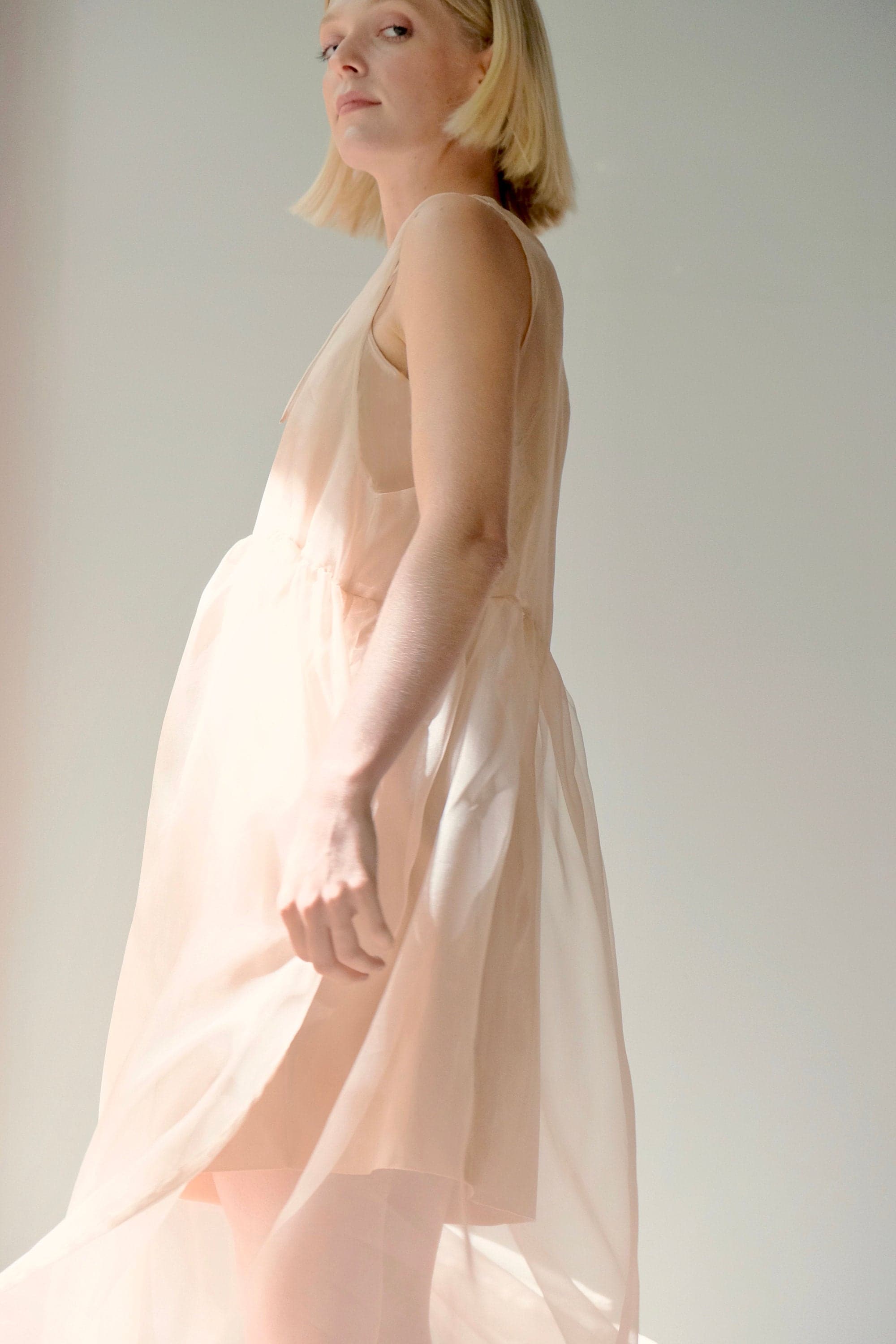 A woman with short blond hair wears a flowing, light peach Coco Silk Dress by Noémiah, expertly crafted from silk organza. She is slightly turned to the side, and the soft lighting enhances the ethereal quality of its relaxed fit against a simple, light-colored background.