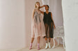 Models pose in the Coco Silk Dress by Noémiah, one in sheer pale pink and the other in black organza, standing beside a decorative white chair. The relaxed fit perfectly complements the soft, neutral background.