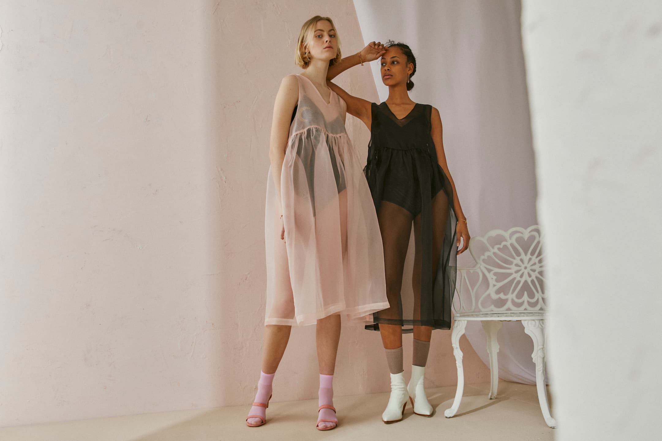 Models pose in the Coco Silk Dress by Noémiah, one in sheer pale pink and the other in black organza, standing beside a decorative white chair. The relaxed fit perfectly complements the soft, neutral background.