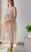 A pregnant woman stands in a well-lit room, wearing a sheer pink Coco Silk Dress by Noémiah and gold shoes. Her light brown hair cascades as she looks ahead. Beside her, a small table showcases a white vase filled with white tulips, adding to the serene scene with its relaxed fit charm.