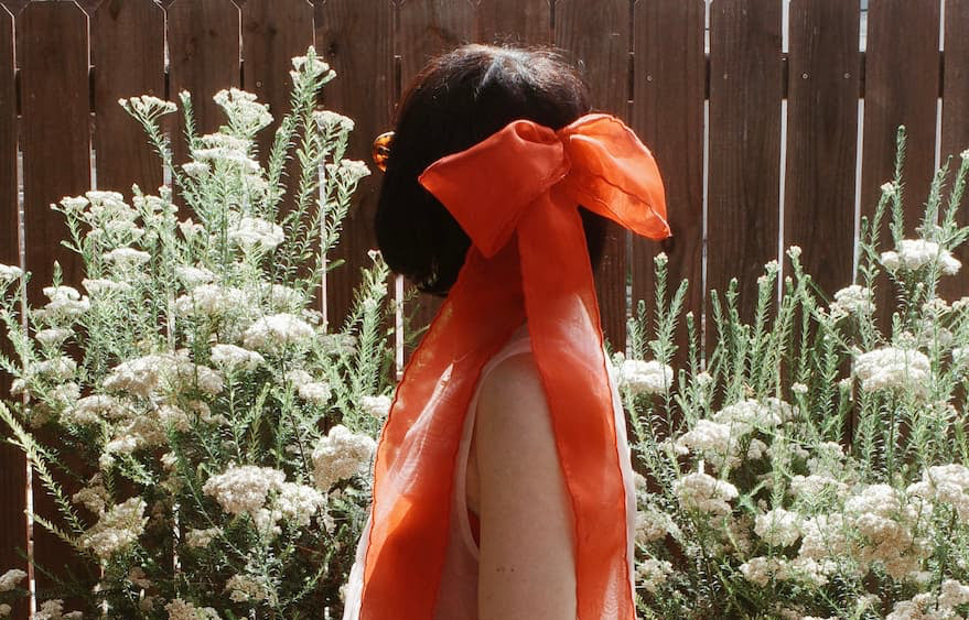 A person with short dark hair faces away, wearing Noémiah's Silk Ribbon — Classic in their hair. They are surrounded by tall white flowers and a wooden fence is visible in the background.