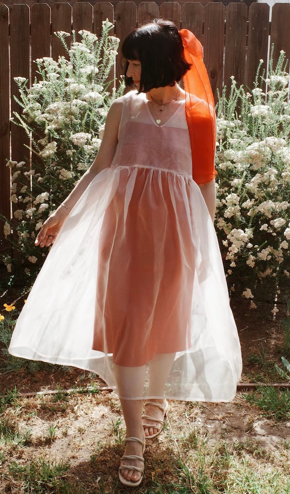 A woman stands on grass among white flowers by a wooden fence, wearing a sleeveless pink dress and sandals. An orange scarf, tied in her hair with the Noémiah Silk Ribbon — Classic, completes her look as she gazes down with one arm out.