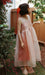 A woman stands in a garden wearing the Noémiah Coco Silk Dress in relaxed pink silk organza with sandals, holding a branch of yellow fruits. She accessorizes with sunglasses and a large red bow in her short dark hair.