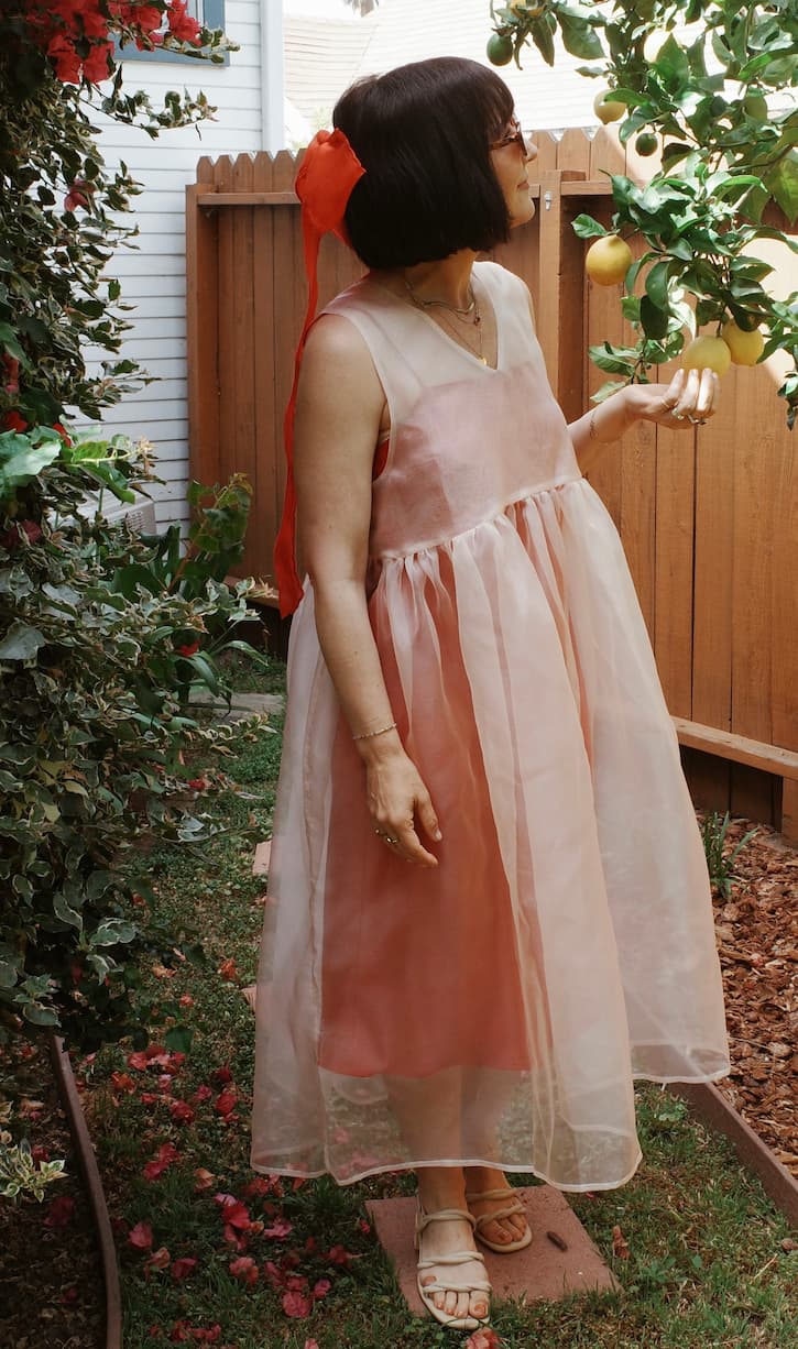 A woman stands in a garden wearing the Noémiah Coco Silk Dress in relaxed pink silk organza with sandals, holding a branch of yellow fruits. She accessorizes with sunglasses and a large red bow in her short dark hair.