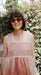 A woman with short dark hair and sunglasses smiles outdoors among green foliage, wearing a relaxed fit sheer pink Coco Silk Dress by Noémiah.