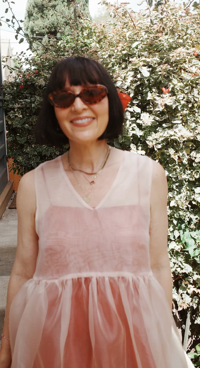 A woman with short dark hair and sunglasses smiles outdoors among green foliage, wearing a relaxed fit sheer pink Coco Silk Dress by Noémiah.