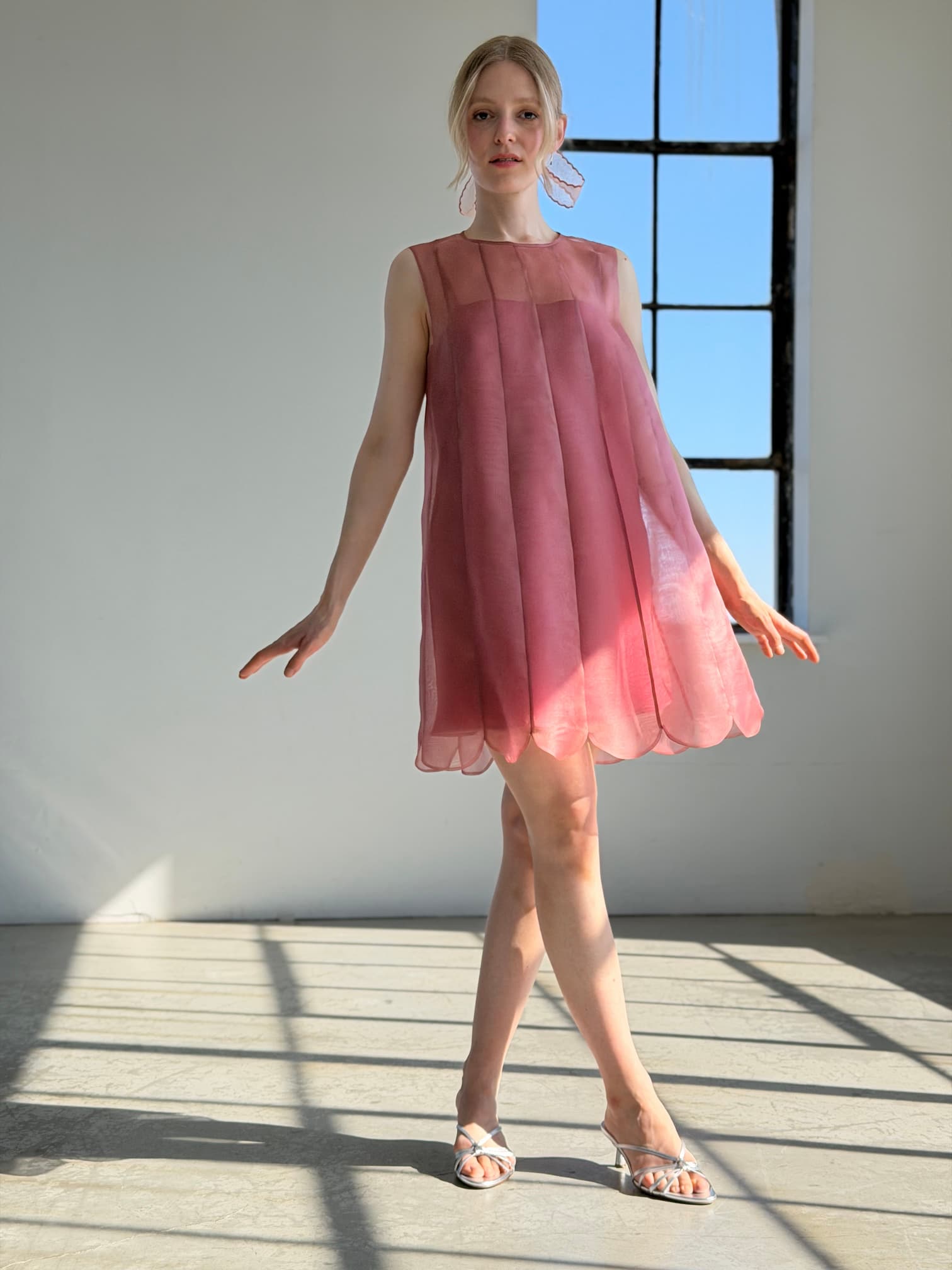 A woman wearing the Daisy Dress by Noémiah, a sleeveless pink organza silk piece, and white heels stands gracefully in a sunlit room by a large window, casting soft shadows on the floor.