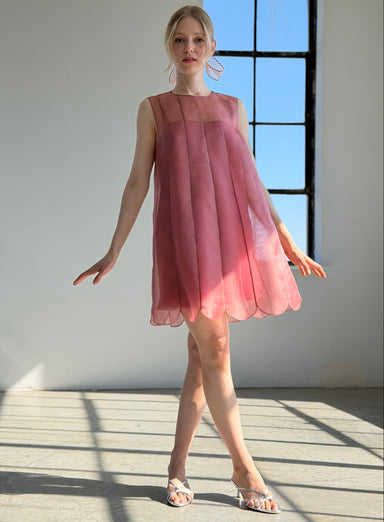 A woman wearing the Daisy Dress by Noémiah, a sleeveless pink organza silk piece, and white heels stands gracefully in a sunlit room by a large window, casting soft shadows on the floor.