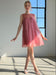A woman wearing the Daisy Dress by Noémiah, a sleeveless pink organza silk piece, and white heels stands gracefully in a sunlit room by a large window, casting soft shadows on the floor.