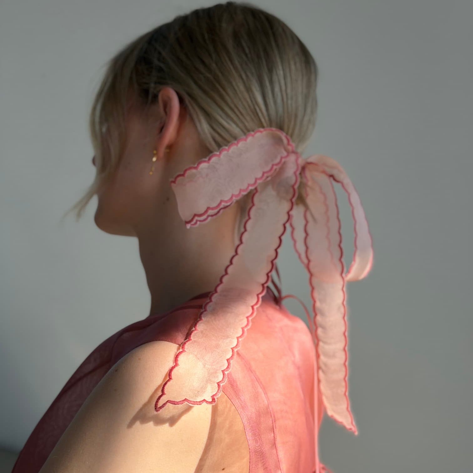 A woman with blonde hair in a low ponytail, tied with a large sheer pink ribbon, wears the sleeveless soft pink Daisy Dress by Noémiah and faces away from the camera.