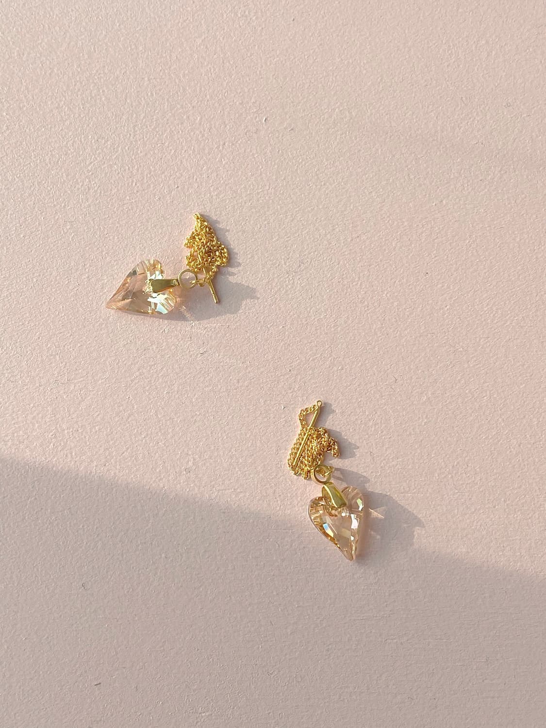 The Danielle Earrings by Noémiah, two pieces adorned with heart-shaped Swarovski crystals and finished in 16K gold plating, rest elegantly on a light surface, their exquisite textured designs above the crystals casting gentle shadows.