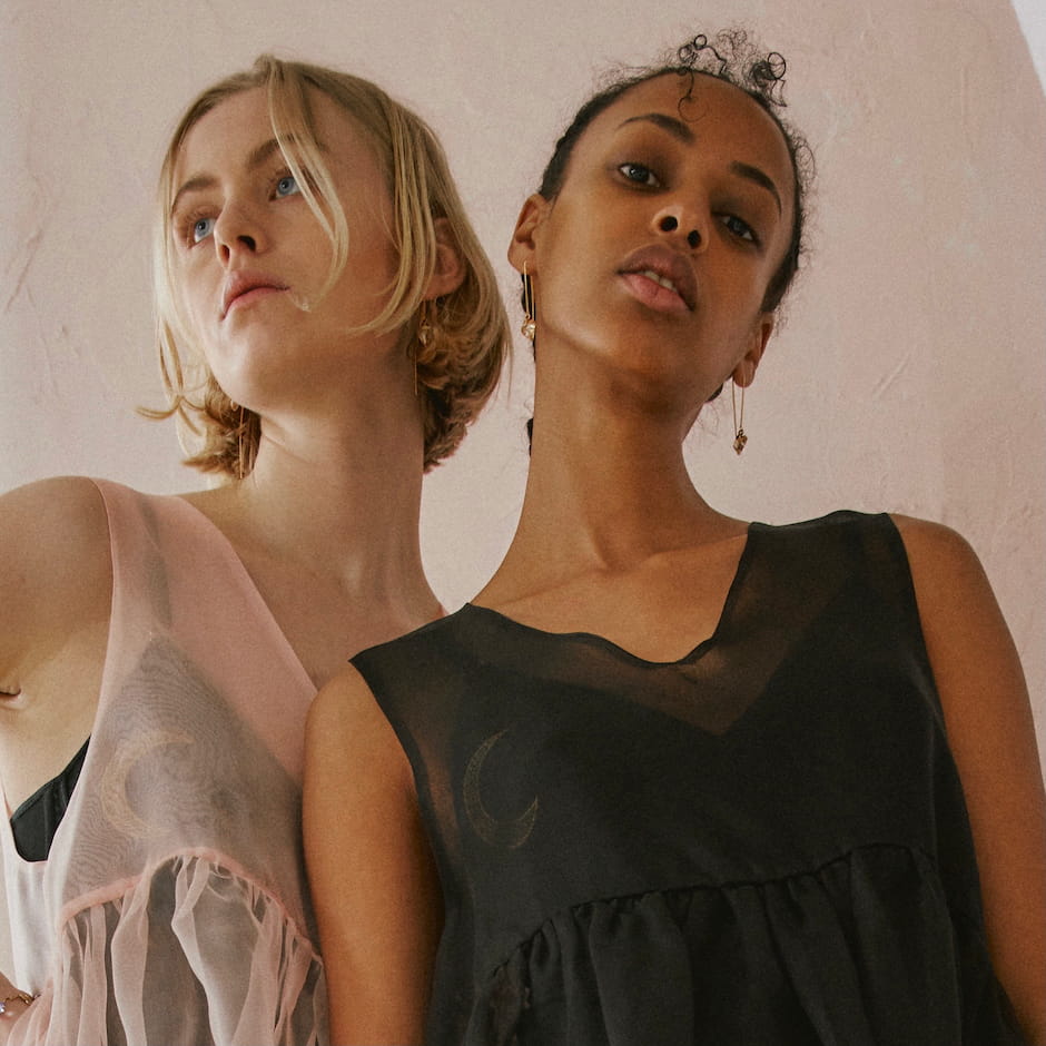 Two individuals stand closely. One wears a light pink dress with ruffles and the stunning Danielle Earrings from Noémiah adorned with Swarovski crystals, while the other dons a sheer black dress featuring a crescent moon design. The soft pink background enhances the serene and stylish atmosphere.