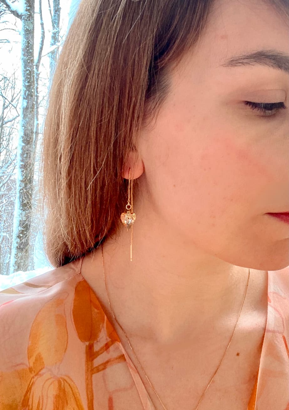 A woman with brown hair is seen in profile, wearing the Danielle Earrings by Noémiah, which are 16K gold-plated and heart-shaped. She also has on a light-colored patterned blouse. The backdrop features a snowy landscape with bare trees.