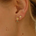 A close-up of an ear showcases two stylish accessories from Noémiah: the Dorothée Earrings. One features a dainty, chic gold flower with a branch-like design, while the smaller one is a simple flower crafted from cubic zirconia. The light skin and few small moles further highlight the delicate beauty.