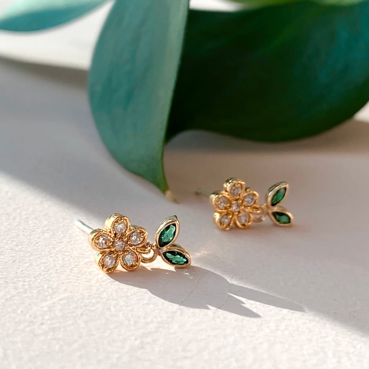 The Dorothée Earrings by Noémiah are gold and diamond floral earrings featuring green leaf accents. They rest on a light surface, partially shaded by a delicate green leaf, offering an elegant touch with dainty chic design and added sparkle from cubic zirconia.