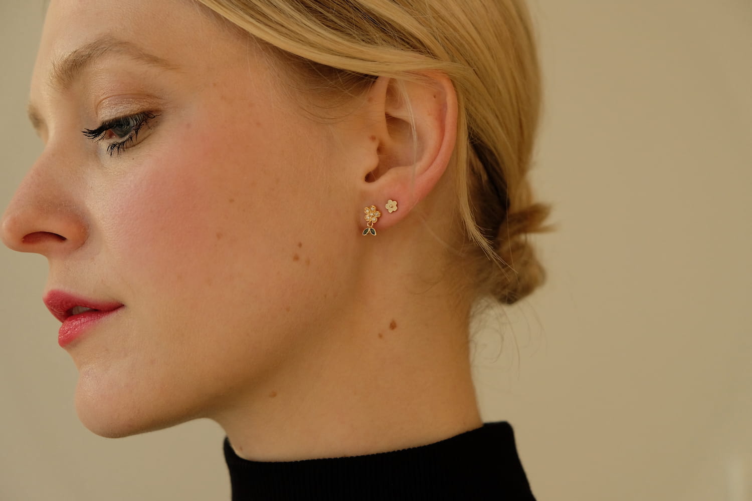 A person with blonde hair is shown in profile against a neutral background, adorned with two elegant Dorothée Earrings by Noémiah: a petite cubic zirconia flower-shaped stud and a bear-shaped stud made from chirurgical steel. They are dressed in a black turtleneck and have light makeup.