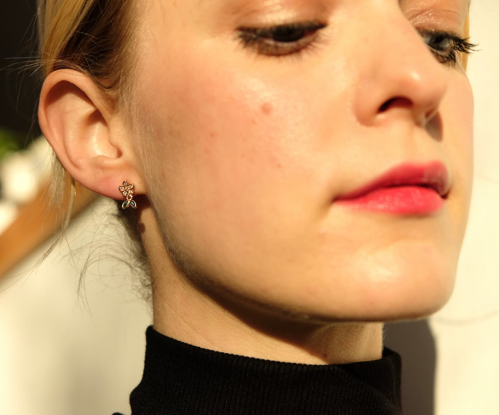 Close-up of a person with light skin wearing a black turtleneck, adorned with the dainty and chic Dorothée Earrings by Noémiah. Their red lipstick complements the warm lighting, which highlights the cubic zirconia set in chirurgical steel along the side of their face and neck.