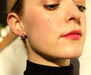 Close-up of a person with light skin wearing a black turtleneck, adorned with the dainty and chic Dorothée Earrings by Noémiah. Their red lipstick complements the warm lighting, which highlights the cubic zirconia set in chirurgical steel along the side of their face and neck.