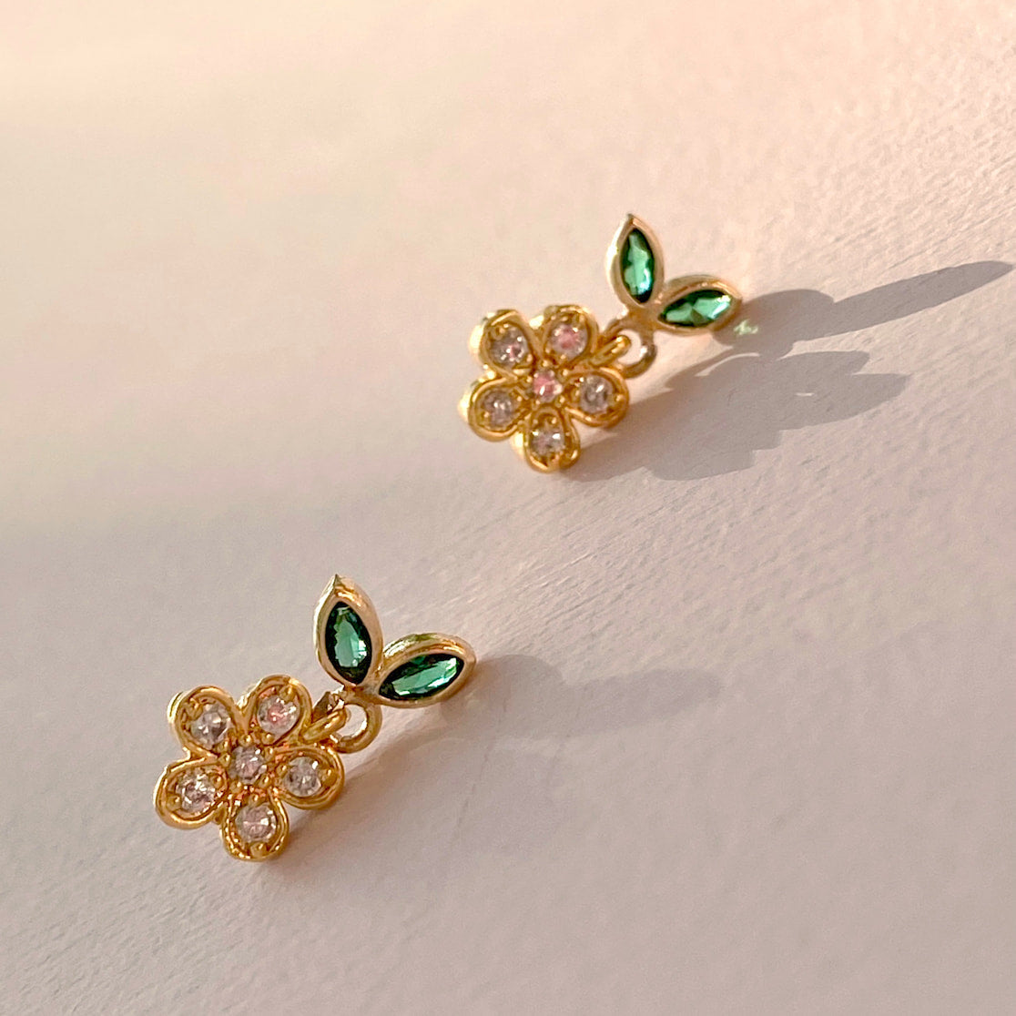 The Dorothée Earrings by Noémiah feature gold flower shapes crafted from chirurgical steel, adorned with clear and pink cubic zirconia petals and accented by green gem leaves. These elegant, stylish earrings are set against a white background, casting gentle shadows.