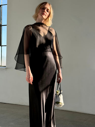 A woman indoors stands in a long black sleeveless dress, draped in the Noémiah Embroidered Shawl, holding a small white floral handbag as sunlight pours through a tall window, casting soft shadows on the wall.