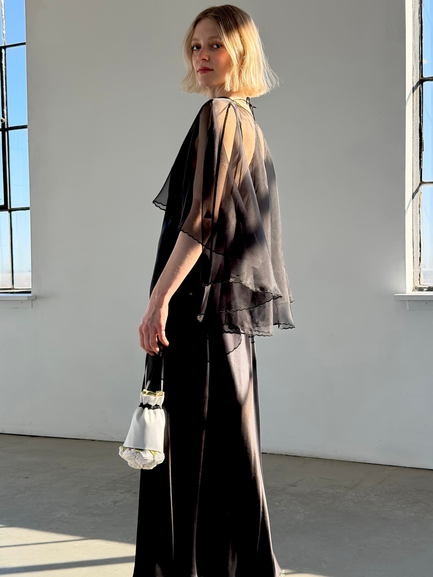 A woman with blonde hair stands in a sunlit room, draped in the Noémiah Embroidered Shawl. She looks over her shoulder as sunlight streams through tall windows behind her, holding a small white handbag.