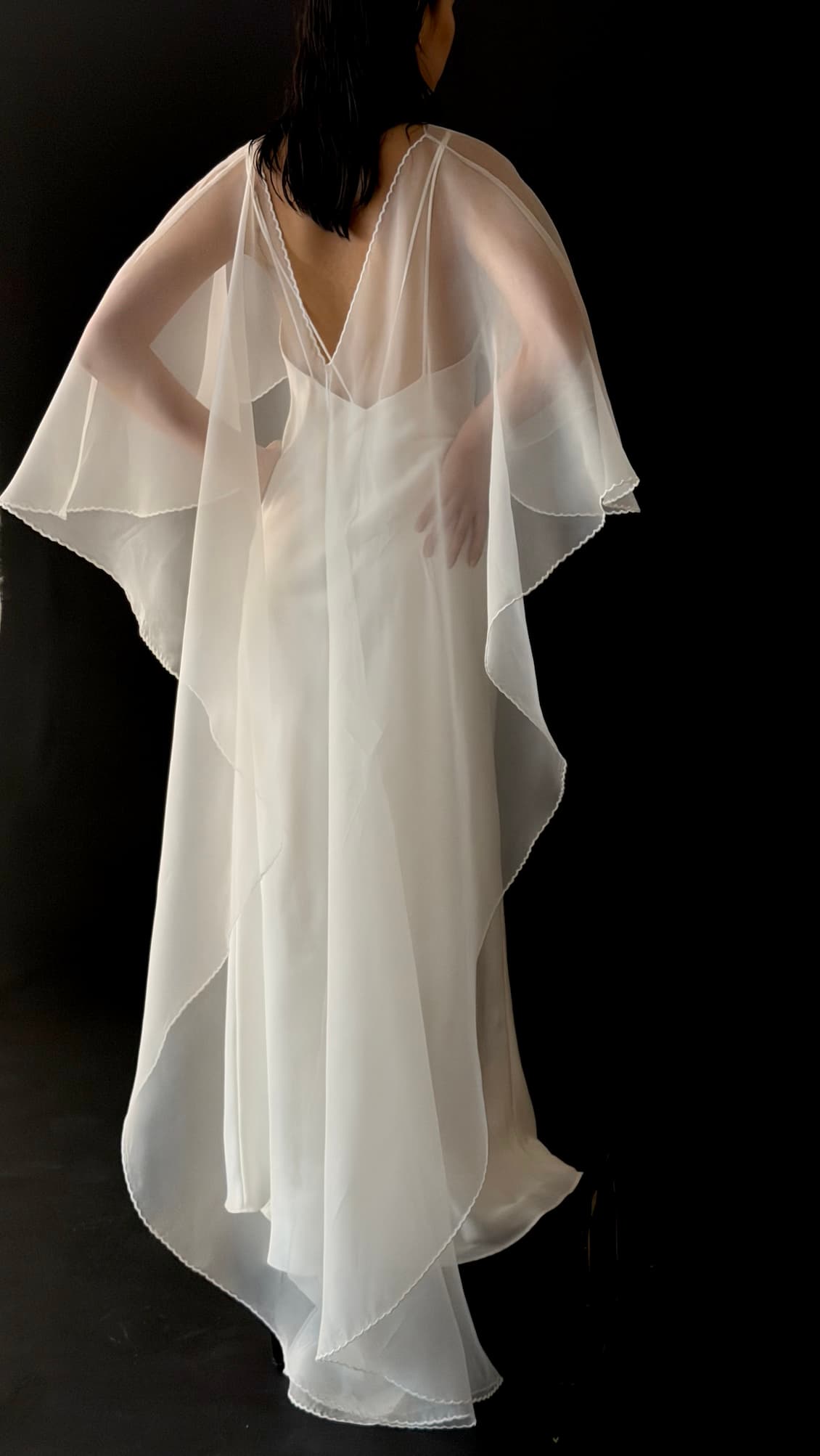 A person poses against a dark background, wearing Noémiah’s Embroidered Shawl over a sheer white dress with wide sleeves and a deep V-back, creating an ethereal and elegant silhouette.
