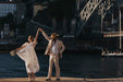 A couple dances joyfully by a river with a large bridge in the background. The woman, wearing Noémiah's Embroidered Tulip Dress, holds a bouquet, while the man wears a light suit and wide-brimmed hat. The sun casts a warm glow over the scene.