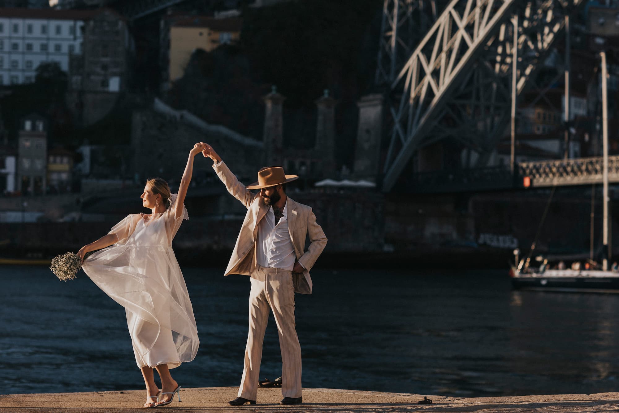 A couple dances joyfully by a river with a large bridge in the background. The woman, wearing Noémiah's Embroidered Tulip Dress, holds a bouquet, while the man wears a light suit and wide-brimmed hat. The sun casts a warm glow over the scene.