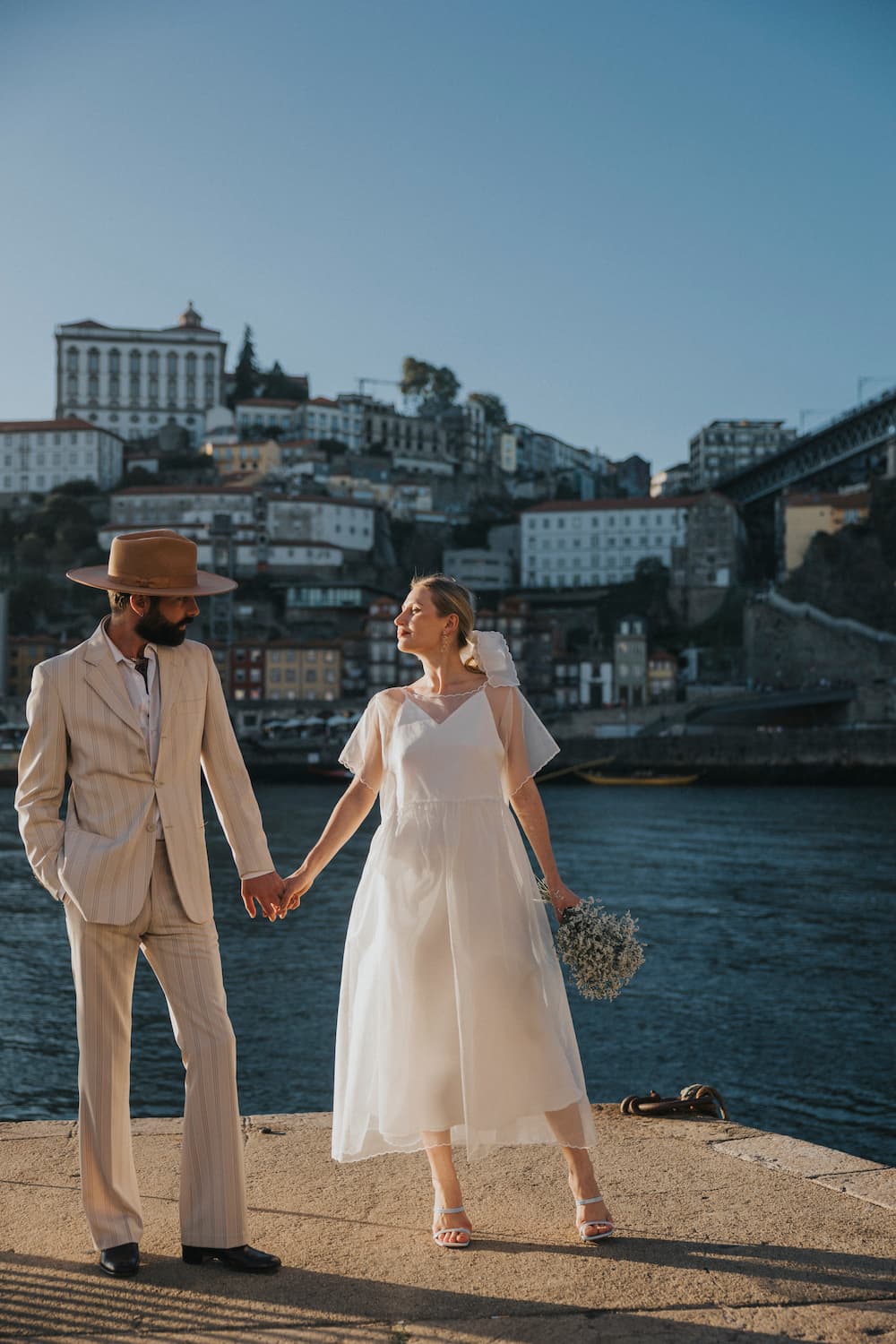 A couple stands hand in hand by the water, with a cityscape in the background. The man wears a beige suit and hat, while the woman is dressed in Noémiah's oversized white Embroidered Tulip Dress made of silk organza, holding flowers. The sun casts a warm glow over the scene, highlighting the romantic atmosphere.