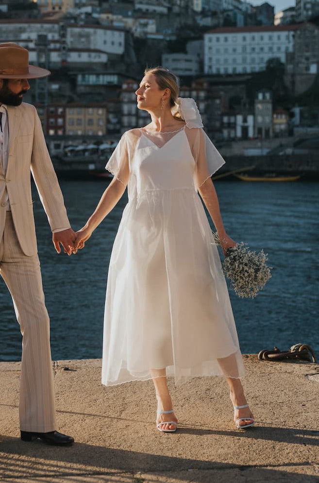 A couple stands hand in hand by the water, with a cityscape in the background. The man wears a beige suit and hat, while the woman is dressed in Noémiah's oversized white Embroidered Tulip Dress made of silk organza, holding flowers. The sun casts a warm glow over the scene, highlighting the romantic atmosphere.