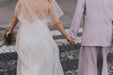 In an enchanting display, a bride in a flowing Embroidered Tulip Dress by Noémiah holds hands with a person wearing a lavender suit. Together, they stroll along a cobblestone street, captured from behind. The bride's small bouquet of flowers elegantly complements the dress's exquisite embroidered finish.