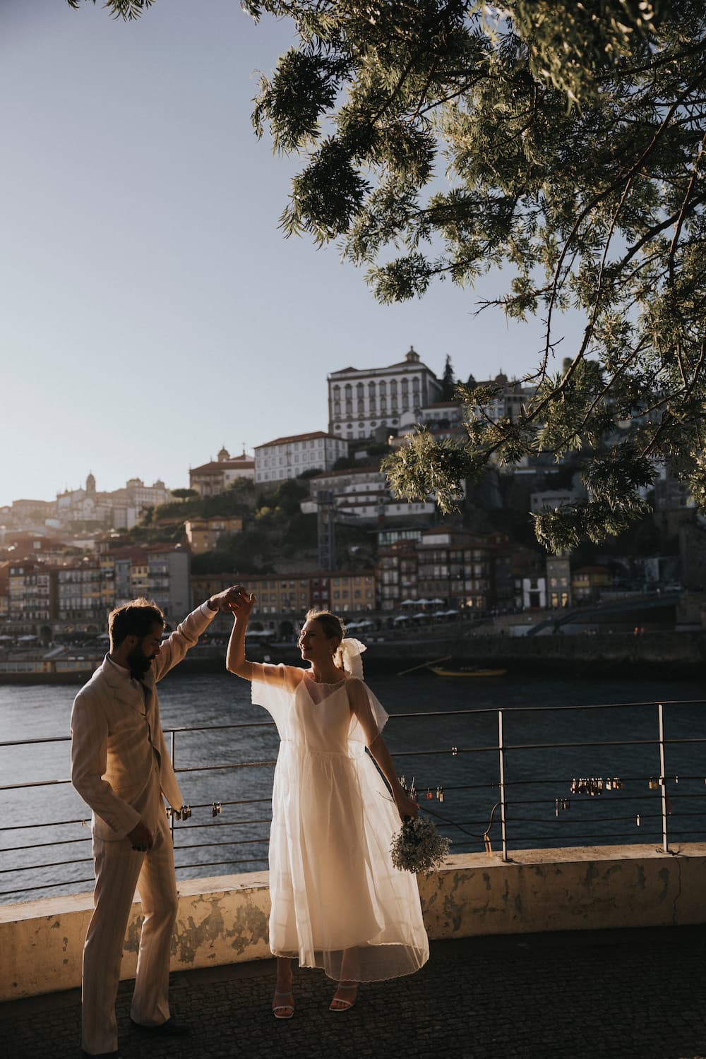 A couple dances joyfully by a riverside railing at sunset, surrounded by lush greenery. The cityscape in the background features historic buildings and a calm river, creating a romantic atmosphere. The bride, wearing the Embroidered Tulip Dress by Noémiah, holds a bouquet.