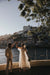 A couple dances joyfully by a riverside railing at sunset, surrounded by lush greenery. The cityscape in the background features historic buildings and a calm river, creating a romantic atmosphere. The bride, wearing the Embroidered Tulip Dress by Noémiah, holds a bouquet.