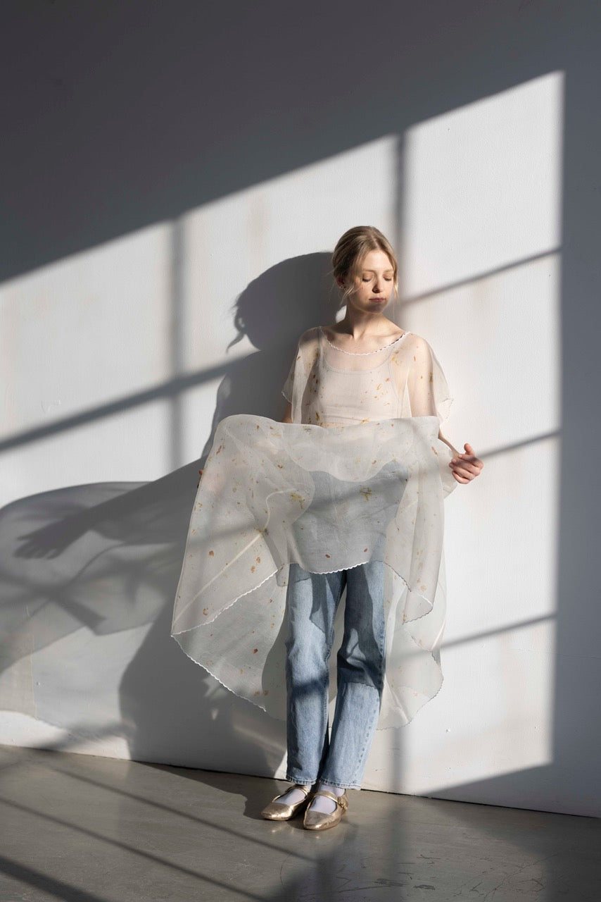 In a sunlit room, a woman is wearing the Embroidered Tulip Dress from Noémiah, styled oversized with an elegant embroidered finish over her jeans and sandals. She gracefully holds out the dress, creating a shadow on the wall behind her. The setting is minimalist, featuring soft lighting and subtle colors.