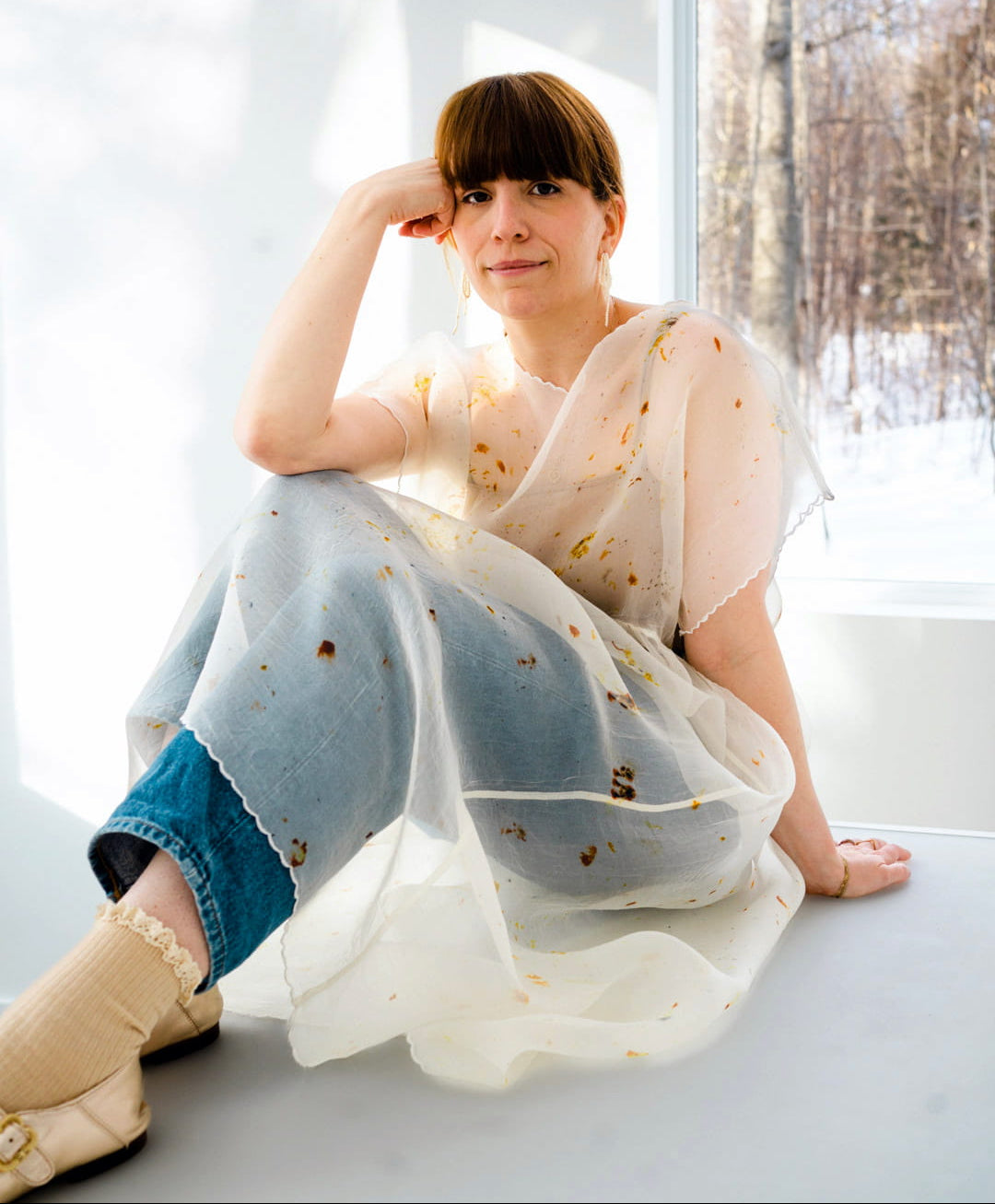 A person with brown hair sits by a window, wearing an oversized Embroidered Tulip Dress from Noémiah over jeans and socks. Sunlight beams in, creating soft shadows. Through the window, snow and trees suggest a winter scene. The individual has a relaxed, thoughtful expression embellished with embroidered details.