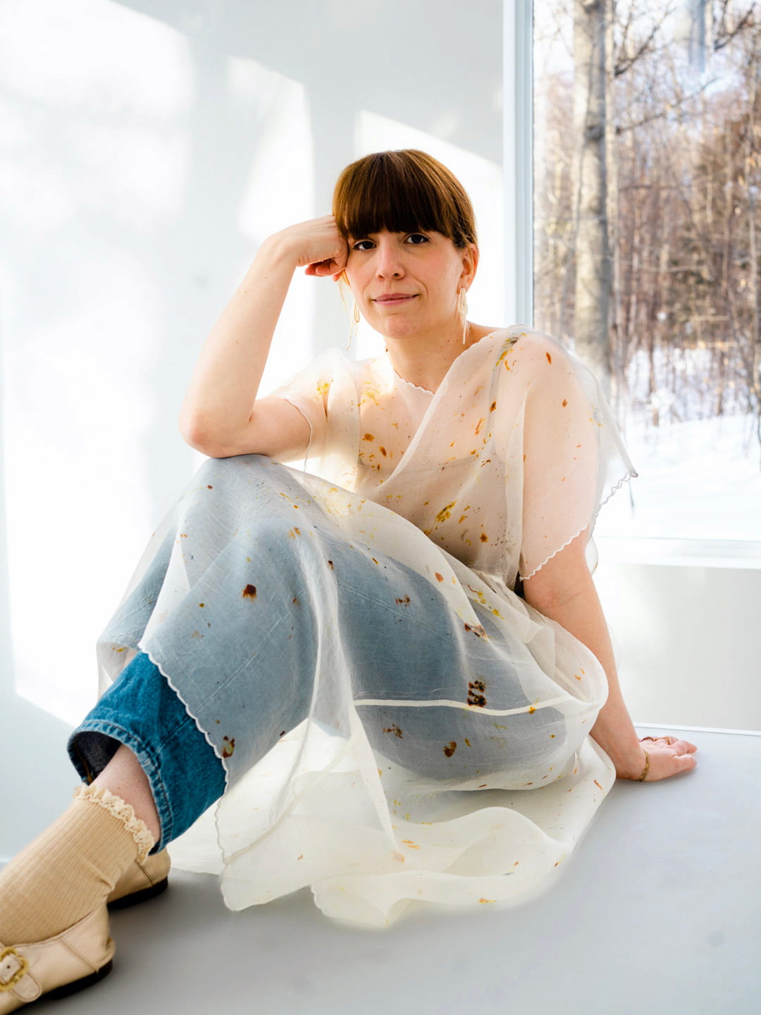 A person with brown hair sits by a window, wearing an oversized Embroidered Tulip Dress from Noémiah over jeans and socks. Sunlight beams in, creating soft shadows. Through the window, snow and trees suggest a winter scene. The individual has a relaxed, thoughtful expression embellished with embroidered details.