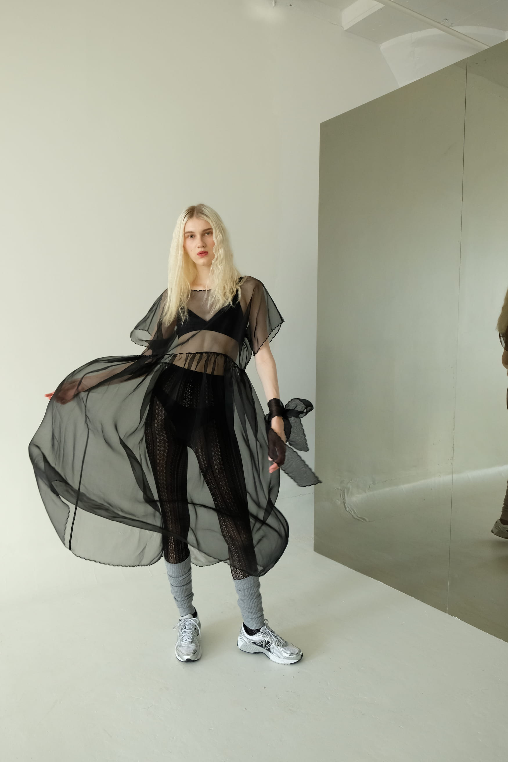 A person with long blond hair is wearing Noémiah's Embroidered Tulip Dress, an oversized black sheer dress, over a crop top and leggings. They have paired it with gray socks and white sneakers. Standing by a large mirror on a light-colored floor, the dress subtly catches the light like silk organza.