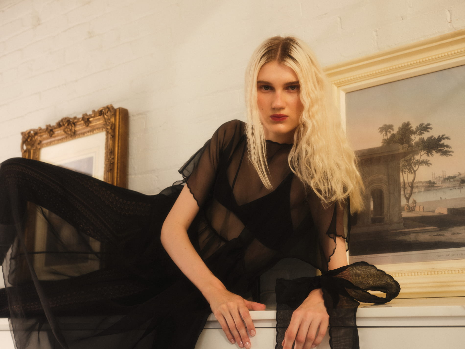 A person with long blonde hair poses elegantly in the Embroidered Tulip Dress by Noémiah, leaning against a white fireplace. Framed paintings hang on the wall in the background, adding an artistic touch to the setting.