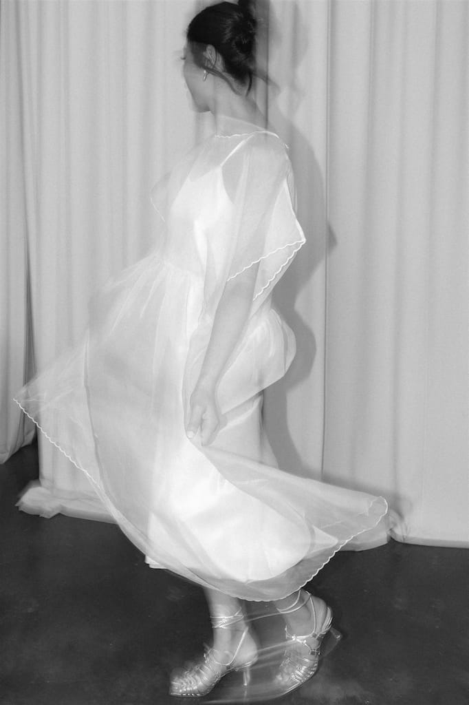 A woman twirls gracefully in a white, flowing Embroidered Tulip Dress by Noémiah, creating a blur effect with her movement. She pairs it with transparent sandals against the backdrop of light-colored curtains. The black and white image highlights the delicate fabric texture and elegant motion.