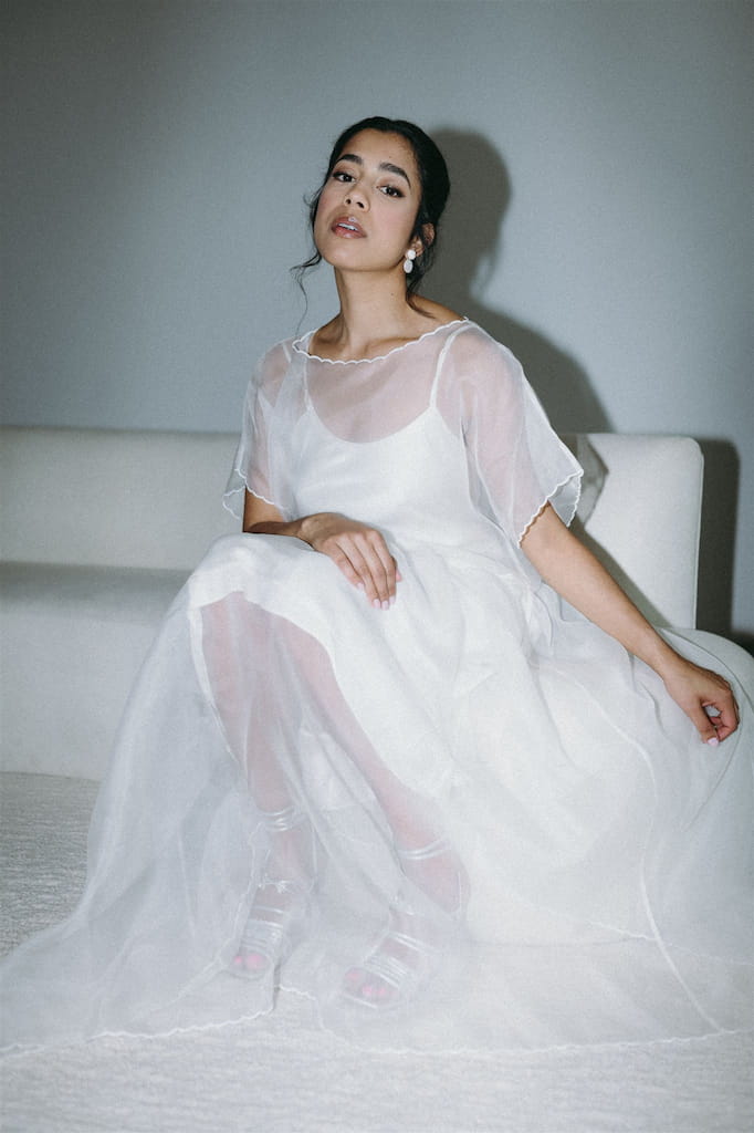 A woman wearing an oversized, sheer white Embroidered Tulip Dress by Noémiah sits gracefully on a white couch against a neutral background. Her hair is styled up, and she gazes slightly upward with a calm expression. The minimalistic setting exudes elegance, enhanced by the dress's subtle embroidered finish.